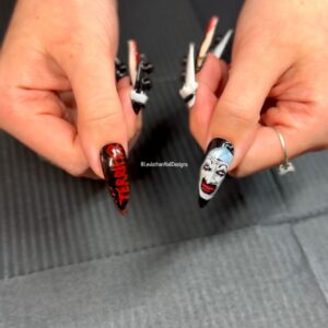 Close-up of two hands with custom horror-themed press-on nails featuring hand-painted 3D details. One nail displays the word "TERRIFIER" in dripping red letters on a black background; the other shows a detailed, creepy clown face with exaggerated makeup and sharp teeth. Set includes long, stiletto-shaped nails with sculptural bone accents.