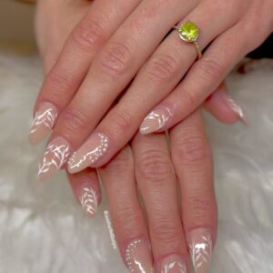 Close-up of elegant almond-shaped press-on nails with a nude base, white hand-painted leaf designs, and pearl accents. A yellow-green gemstone ring is worn on the ring finger, with the hands placed on a soft white surface.