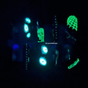 Two images of custom press-on nails: one in bright light showing sculpted gothic horror nails with eyeballs, skulls, webs, barbed wire, and red gore detailing; the other in the dark showing neon green and blue glow-in-the-dark effects on the same nails.