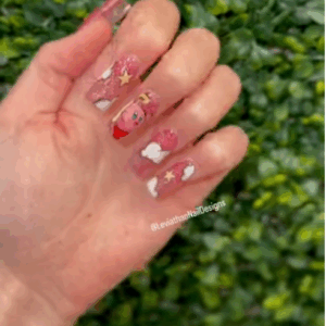 Long coffin-shaped glitter pink press-on nails with hand-painted Kirby character, white clouds, yellow stars, and sculpted 3D accents. Set shown against a leafy green background.