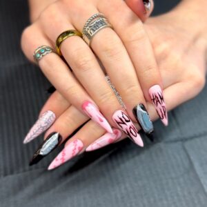 Long stiletto press-on nails with a matte pink base and black accents, featuring hand-painted text (“NO,” “HANDS OFF”), dripping designs, a sculpted silver knife charm, and spiderweb detailing.