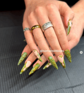 Long stiletto press-on nails with sage green and nude matte base colors, decorated with sculpted golden vines. Nails shown on crossed hands with ornate silver and gold rings, photographed on a dark surface.