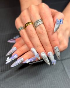 Close-up of long, sculpted press-on nails with a metallic lavender and silver base. Each thumb nail is shaped like an angel wing and decorated with sparkling 3D butterfly gems in blue and violet.